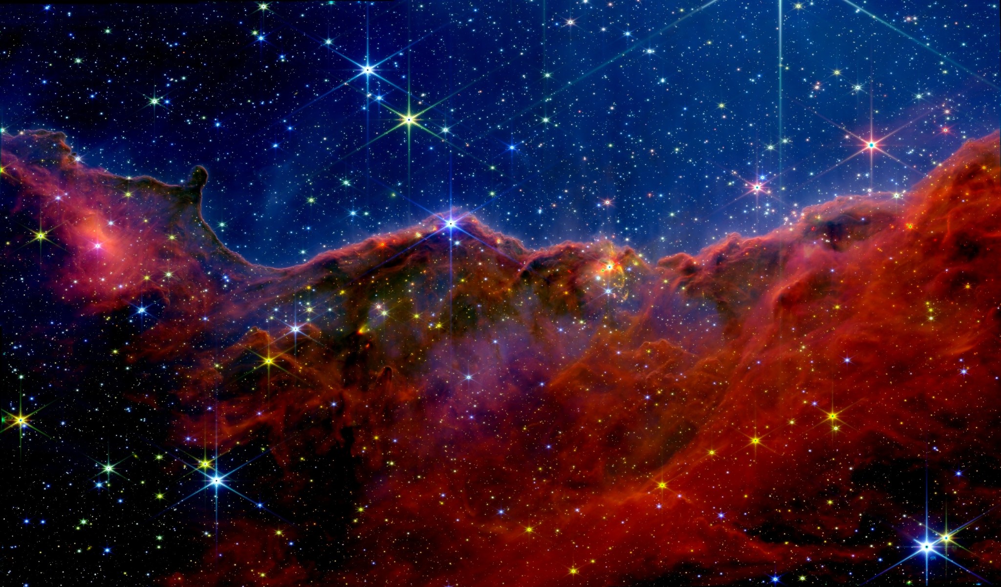 The Cosmic Cliffs in NGC 3324 — a JWST NIRCam image reprocessed by Kyle Eberly. A rugged, mountain-like ridge of orange-brown nebular dust runs across the lower half, studded with stars showing JWST's six-point diffraction spikes, beneath a deep-blue sky full of stars.